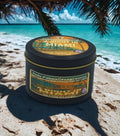Candle tin labeled 'A Night in Miami' on a beach with palm trees and ocean in the background