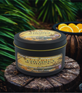 Tin of 'Citrus Coconut Mahogany' candle on a wooden surface with greenery in the background
