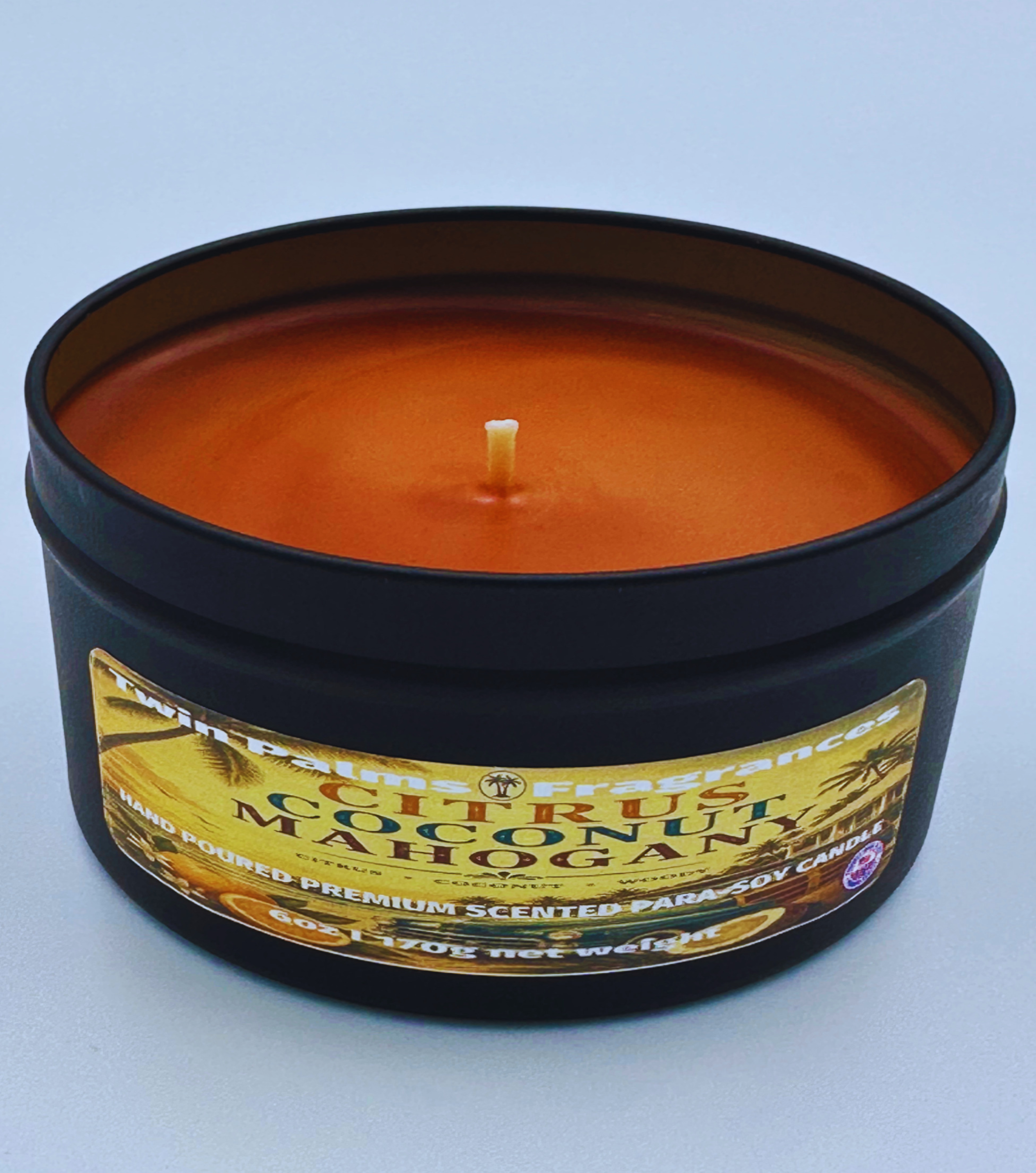 Citrus Coconut Mahogany  Scented candle in a black tin with a label on a light blue background