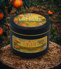 Candle labeled 'Peach Orchard' by Twin Palms Fragrances on a wooden surface with peach tree in the background.